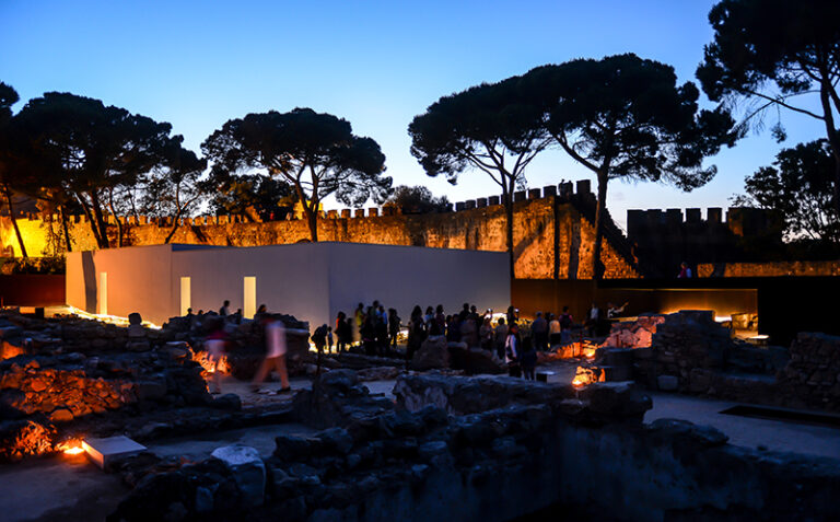 Descrição da imagem: fotografia do Núcleo Arqueológico tirada ao entardecer. O céu começa a escurecer, as luzes do Núcleo Arqueológico estão acesas, e, no centro da imagem, veem-se pessoas a circular perto das casas islâmicas.
