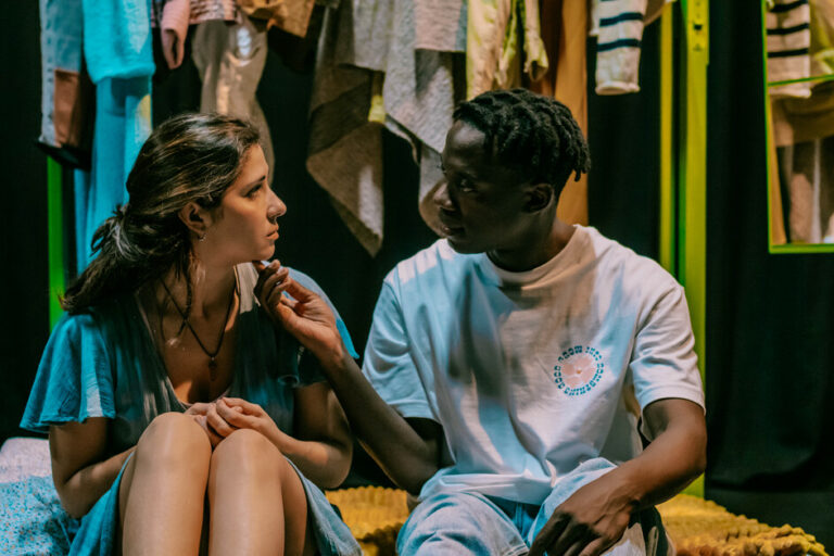 dois jovens sentados na cama, vestidos olham um para o outro