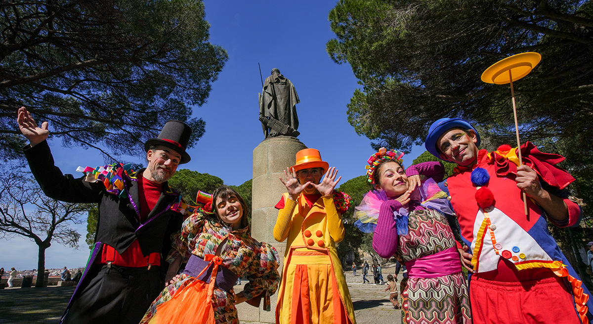 Descrição da imagem: fotografia dos animadores do Chapitô, vestidos com roupas coloridas, na Praça d'Armas do Castelo de São Jorge, com a estátua de Dom Afonso Henriques por trás.
